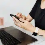 Person using a smartphone while working on a laptop at a desk